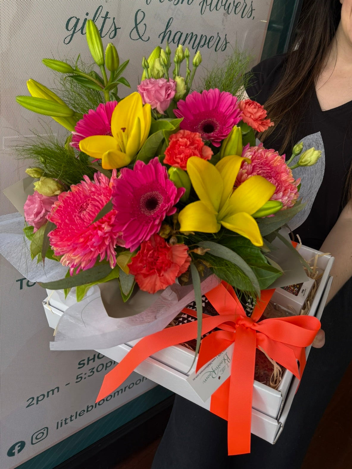 Woman holding a colorful bouquet of flowers with an 'Exquisite fresh flowers, gifts & hampers' sign in the background.